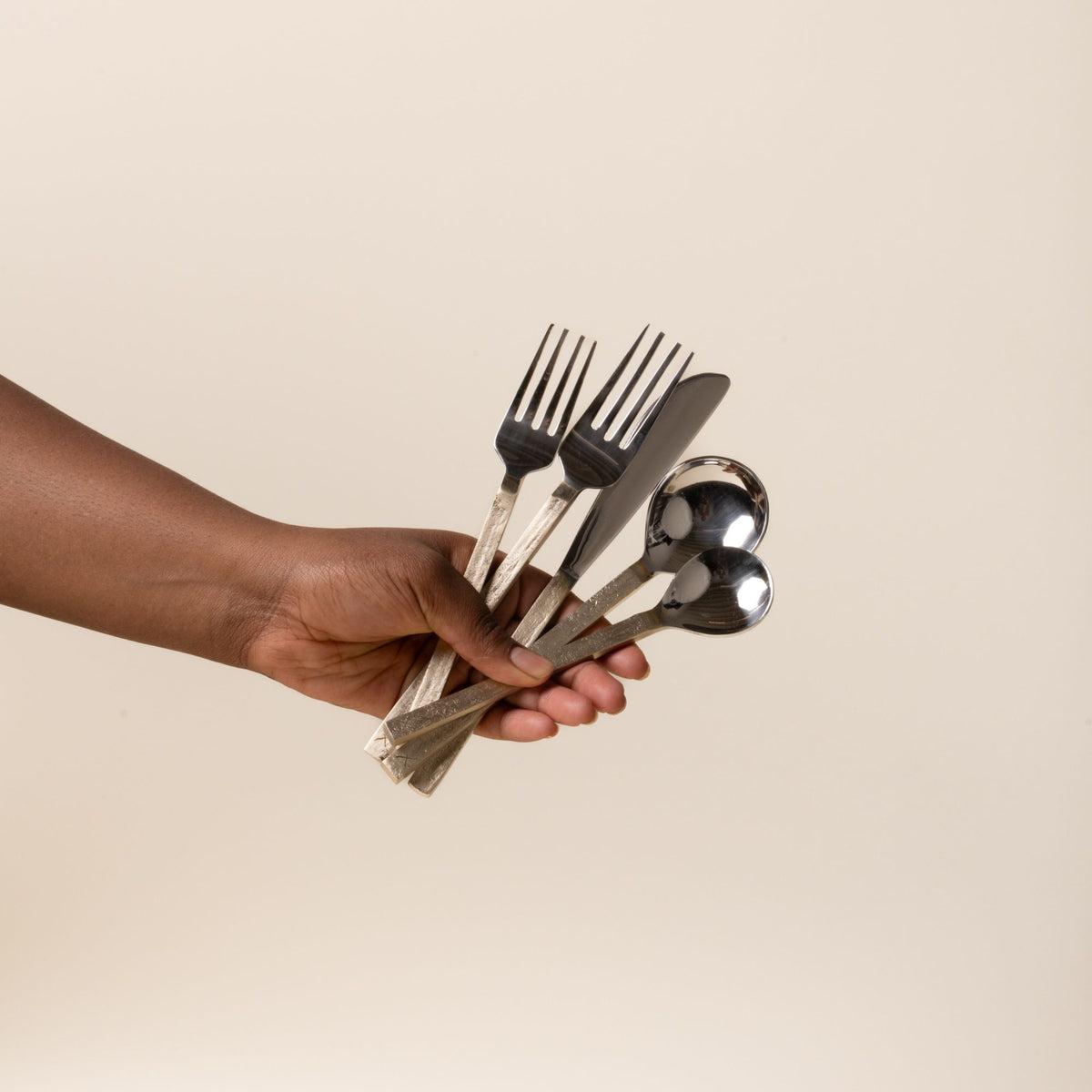 A hand holds out five-piece stainless steel and brass flatware set with two forks, a knife, and two spoons against a light background