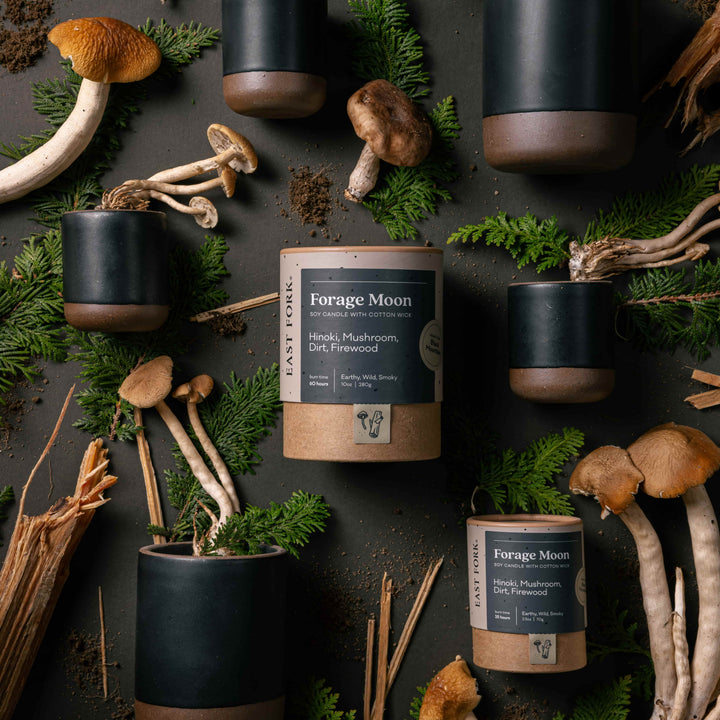A cylindrical ceramic vessel in an muted charcoal color laying on its side - mushroom, greenery, and firewood are artfully styled coming out of the top of the candle to reflect the scent.