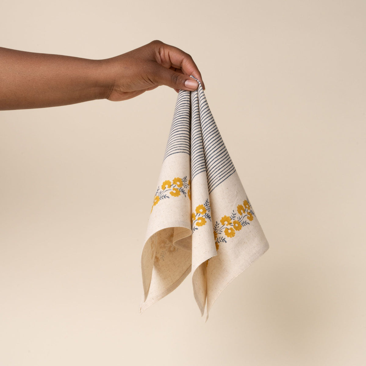 A hand holds out a cream-colored napkin with a delicate yellow floral border framing a central blue striped patchwork design, against a neutral background.