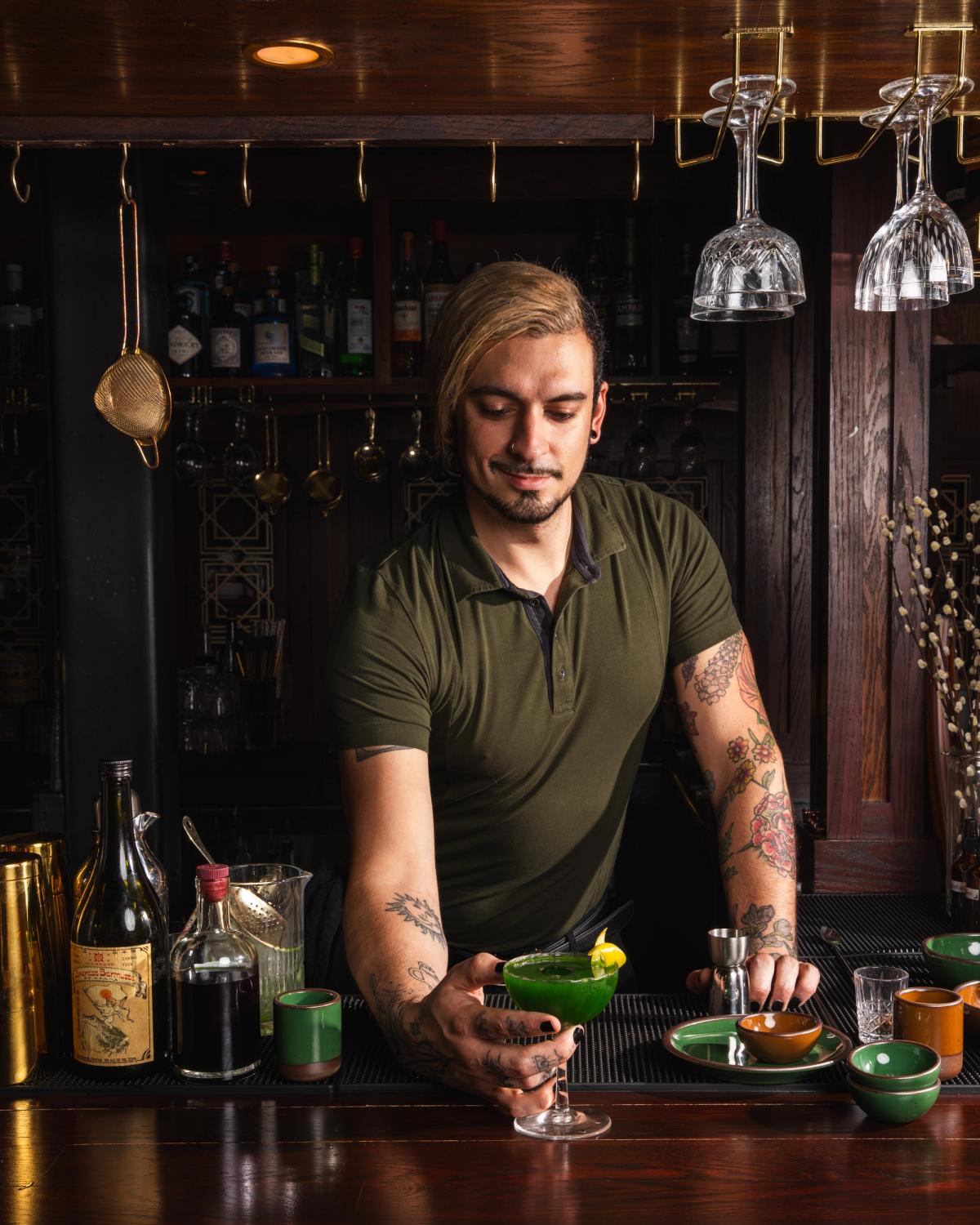 A bartender stands behind a dark wooden bar and hands over a green cocktail in a stemmed glass.
