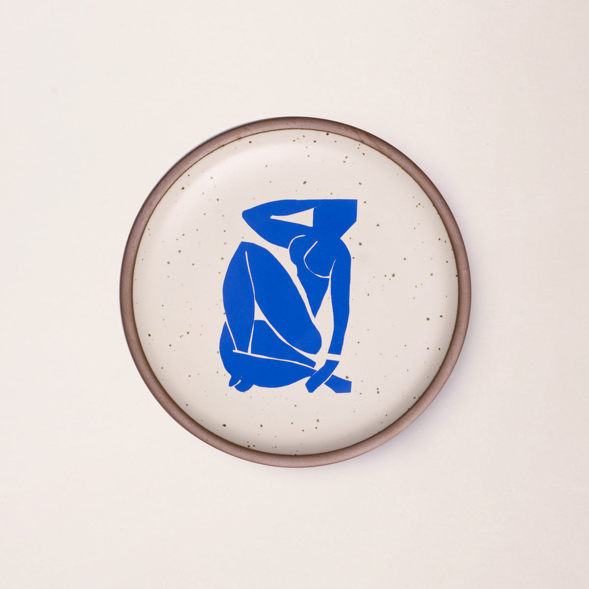 A plate featuring Nu Bleu III, by Henri Matisse, a bright blue, abstract, silhouetted female figure in a curled, seated position, with sharp cut-out edges, centered on an off-white ceramic plate with an unglazed rim.