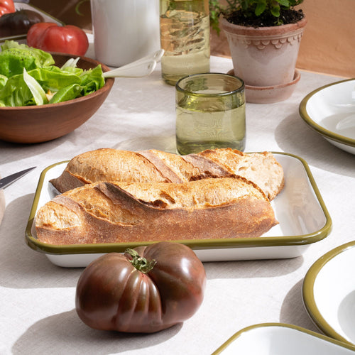 On a tablescape, there are 2 short baguettes on a large rectangular baking dish in a white color with an olive green rim.