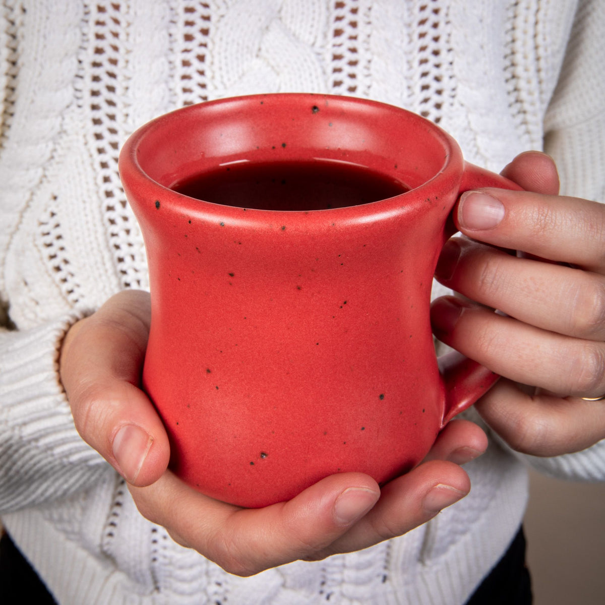 A hand holds a classic diner mug that tapers out on the top and bottom in a bold red color featuring iron speckles and a handle.
