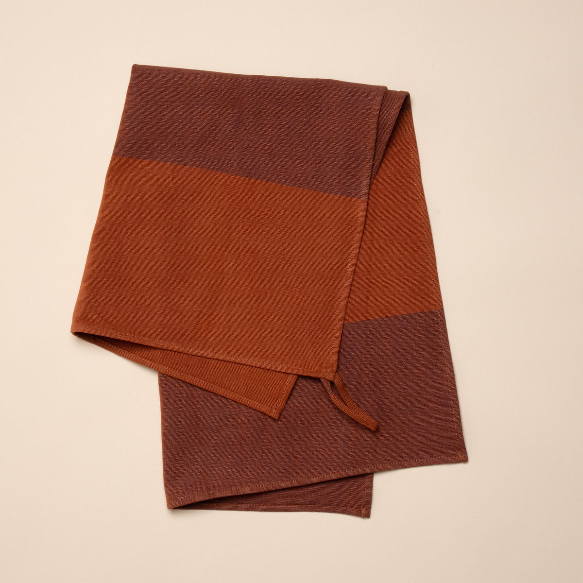 A folded tea towel with color blocks of rust and dark red, and featuring a hanging loop on a corner