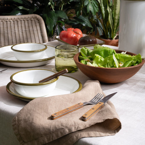 A tabletop filled with white enamel plate and bowl table setting with flatware with wood handles, along with a napkin, wood bowl, and glass.
