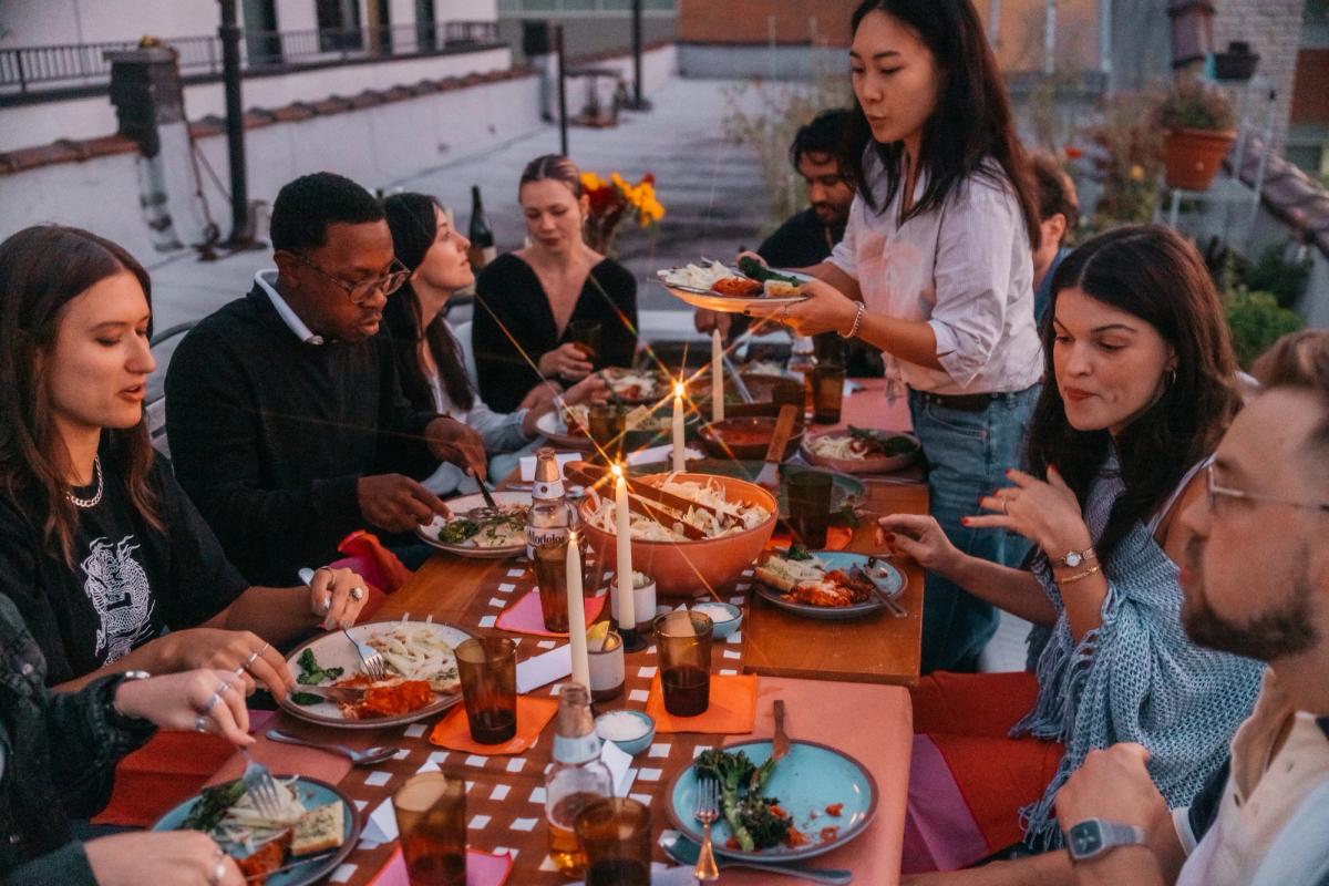 A group of people sit around a dinner table set with food and candles outside at dusk.