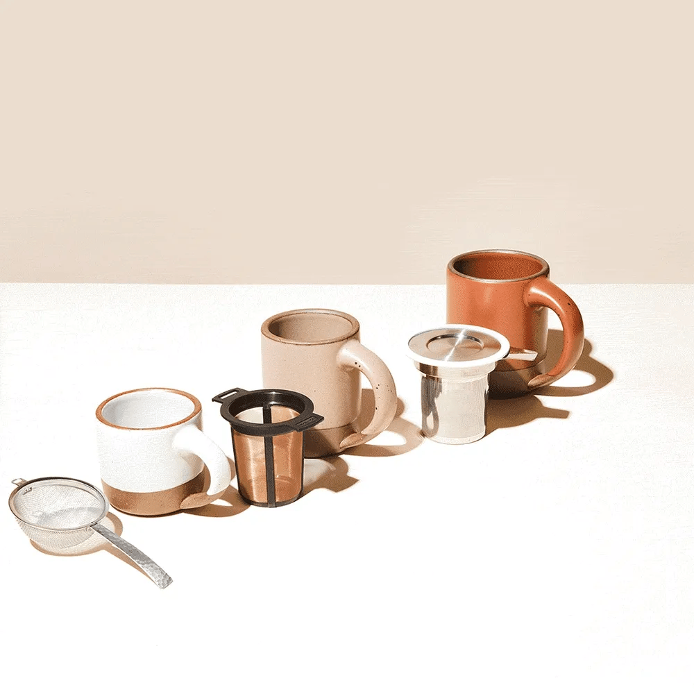 Three ceramic mugs in varying earth tones with infusers and a metal tea strainer are arranged on a light surface against a beige background.