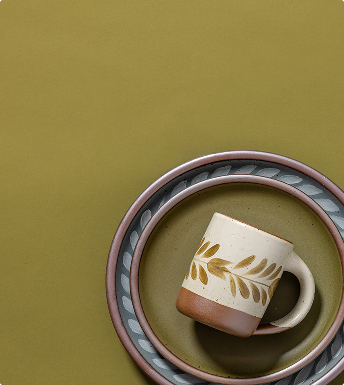A ceramic mug with a leaf pattern on a cream and brown body, styled on a matching patterned plate against a solid olive green background.