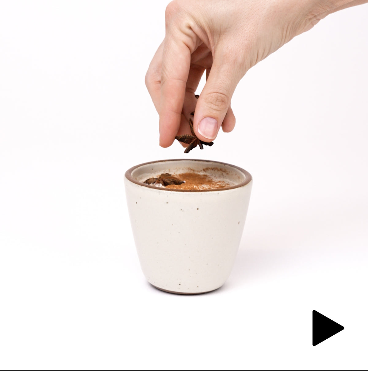 A video of a hand setting down a tapered cup filled with a latte and another hand sprinkling star anise on top.