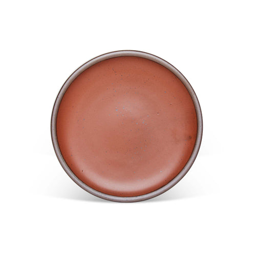 A dinner sized ceramic plate in a cool terracotta color featuring iron speckles and an unglazed rim.